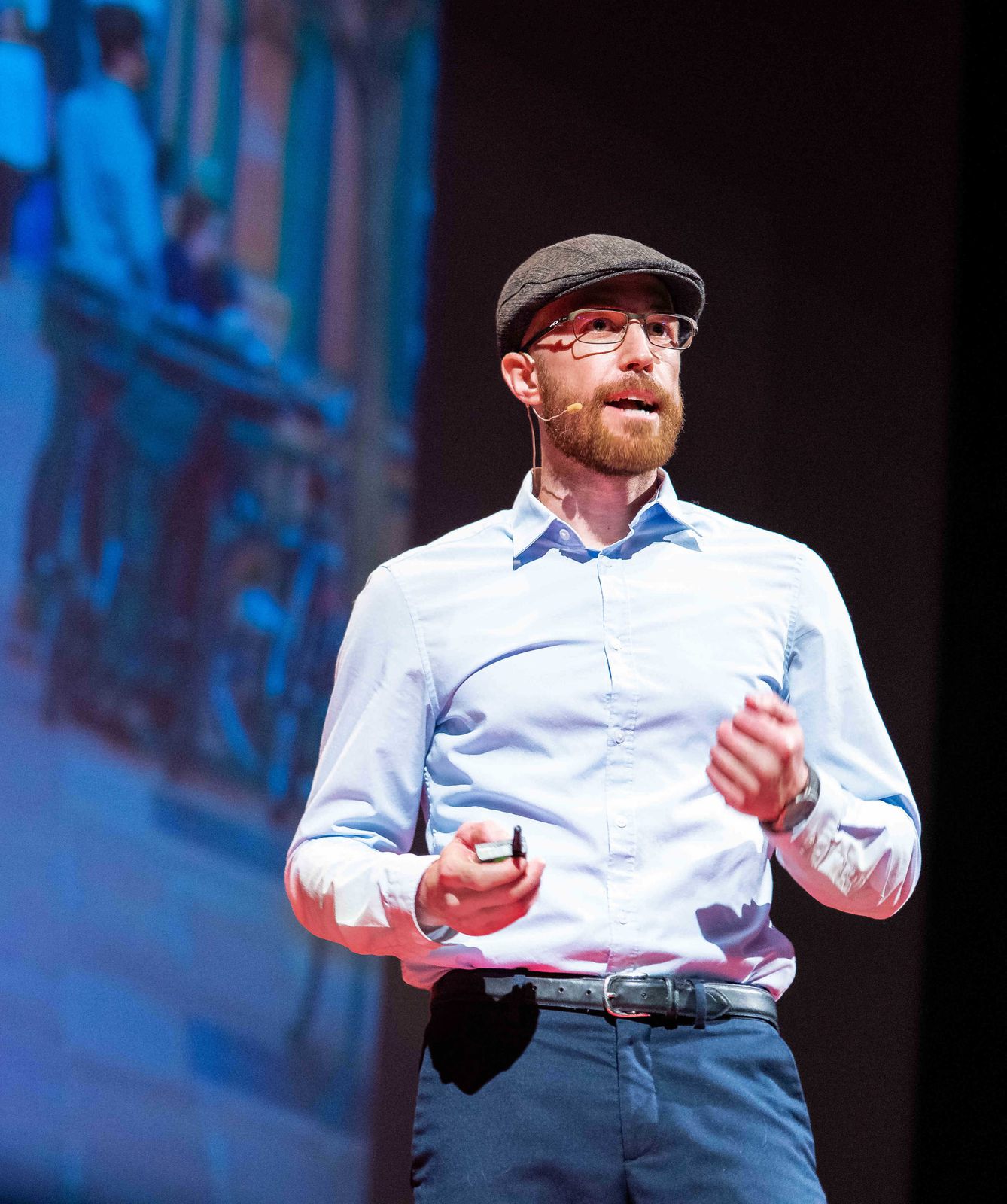 Justin Lewis speaking on stage at TEDxGrandJunction, wearing a flat cap and glasses