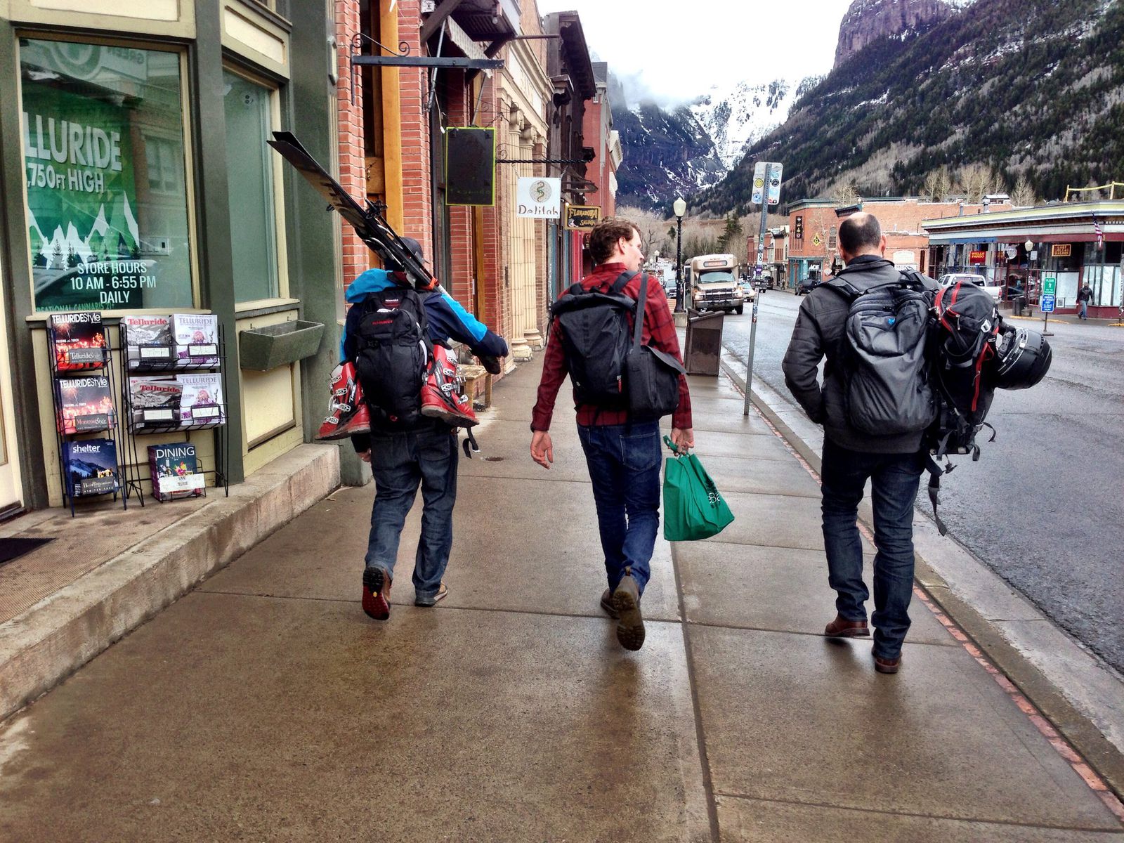 Three people carrying backpacks and ski gear walk down a mountain-town main street toward snowy peaks
